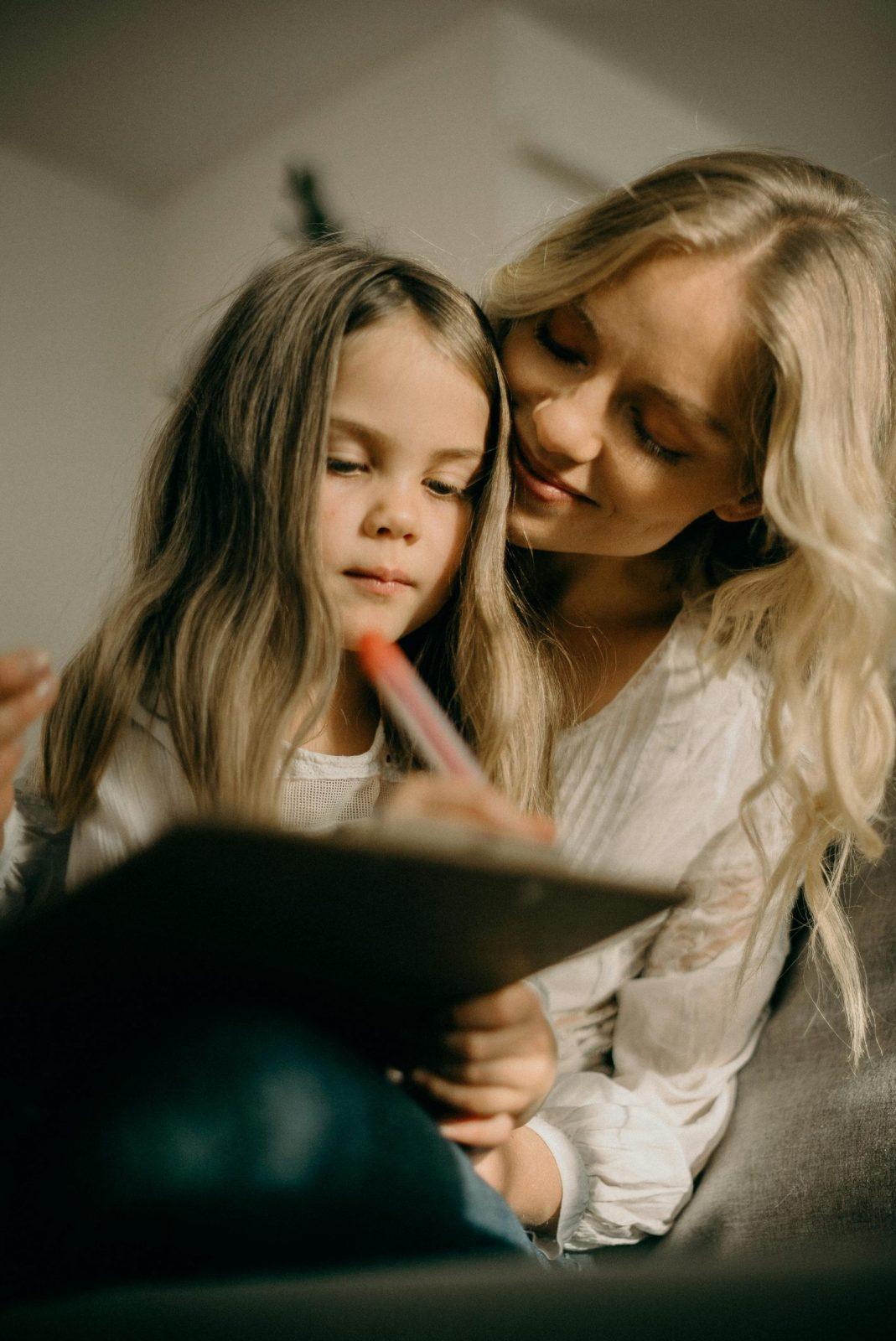 A blonde woman looking over the shoulder of a a young blonde girl writing on a piece of paper