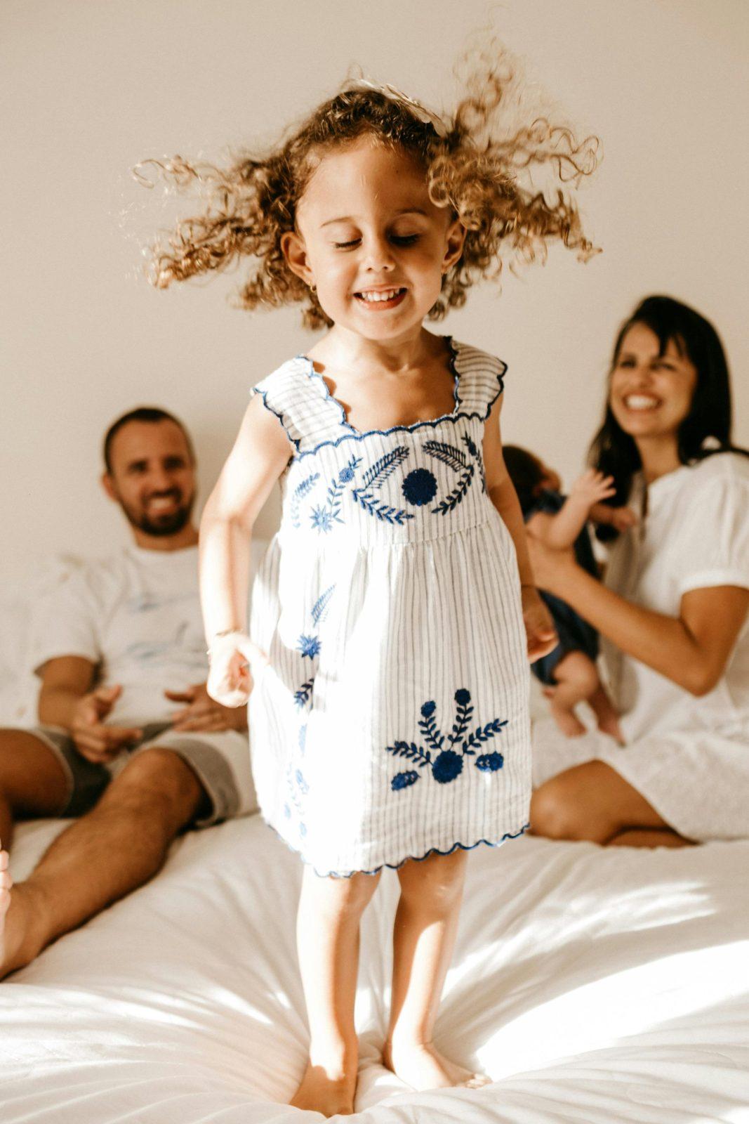 A young girl jumping on a bed with her parents smiling in the background